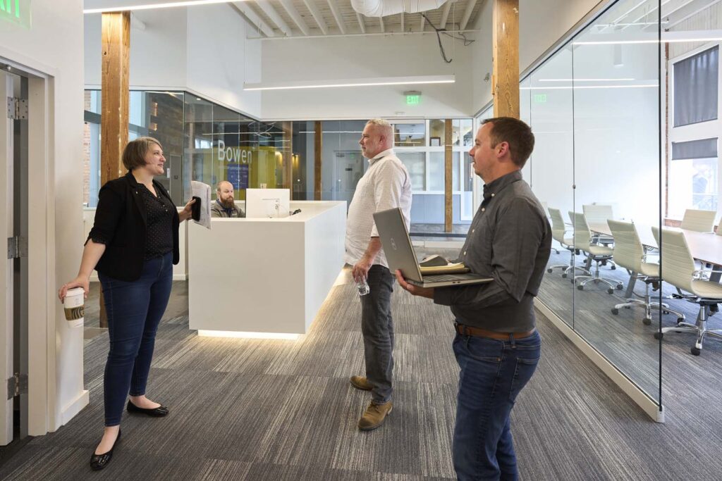3 people talking in front of a reception desk while others are meeting in a glass conference room in the background